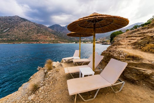 Summer vacation destination. Straw sunshades and sunbeds on the empty pebble beach with sea in the background. Vacation And Tourism Concept. Sunbeds On The Paradise Beach. Umbrellas and sunbeds.