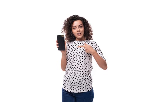 young stylish woman with curly hair dressed in a blouse with polka dots shows a smartphone with a mockup for advertising.