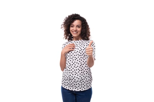 charming young brunette curly woman dressed in summer blouse with pea print points with index finger.