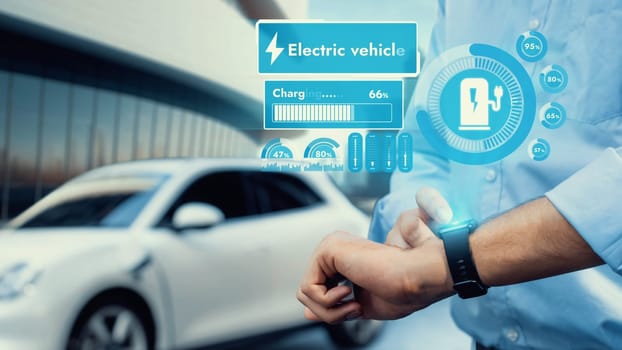 Businessman check EV car battery status on smartwatch hologram while recharge from charging station with modern building background. Futuristic lifestyle of clean energy for EV tech adaptation. Peruse