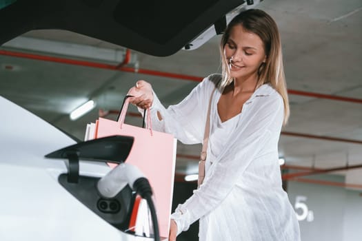 Young woman travel with EV electric car to shopping center parking lot charging in downtown city showing urban sustainability lifestyle by green clean rechargeable energy of electric vehicle innards