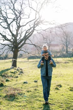 Dad in sunglasses with a little girl on his shoulders walks through a sunny pasture against the backdrop of mountains. High quality photo