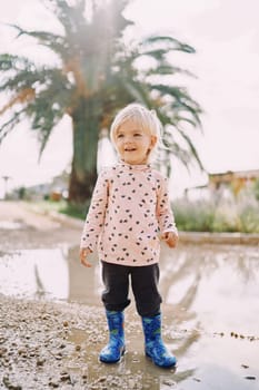 Smiling little girl in rubber boots stands in a puddle against the background of a green palm tree. High quality photo