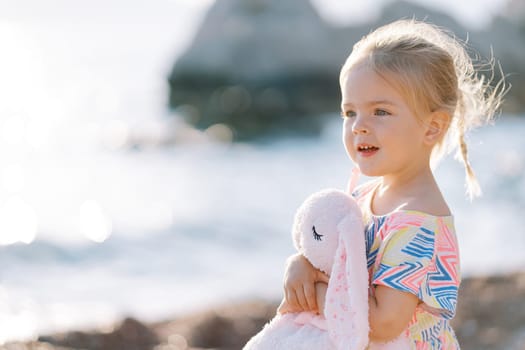 Little surprised girl stands on the beach hugging a toy hare. High quality photo