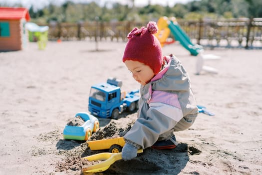 Little girl squatting puts sand with a toy shovel on a plastic car. High quality photo