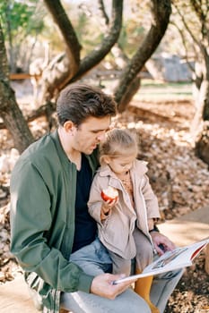 Dad reads a book to a little girl with an apple sitting on his lap on a bench in the garden. High quality photo