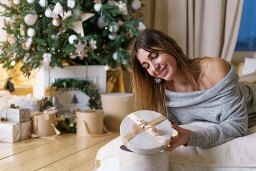 Beautiful caucasian woman lying on bed with gift in her hands, looking and smiling against background of a christmas tree. A festive evening in bed is a happy girl. Cozy photo of the new year