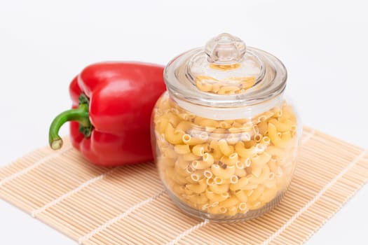 Uncooked Chifferi Rigati Pasta in Glass Jar and Red Bell Pepper on Bamboo Mat on White Background. Fat and Unhealthy Food. Classic Dry Macaroni. Italian Culture and Cuisine. Raw Pasta