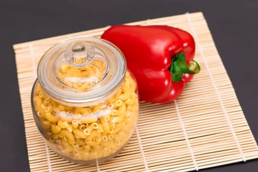 Uncooked Chifferi Rigati Pasta in Glass Jar with Red Bell Pepper on Bamboo Mat on Black Background. Fat and Unhealthy Food. Scattered Classic Dry Macaroni. Italian Culture and Cuisine. Raw Pasta