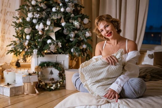 Beautiful happy woman in white shirt sits on bed covered by pillow in room with christmas tree. Seductive caucasian girl with makeup and red lipstick is sitting comfortably in evening on eve holiday