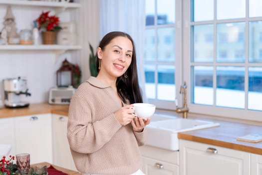 Concept festive Christmas atmosphere, cute woman drinking tea or coffee in a decorated kitchen wearing a sweater