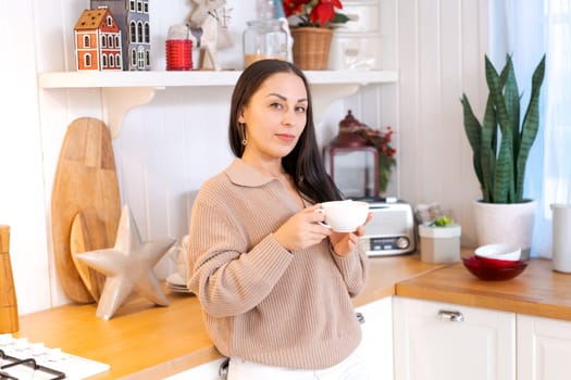 Concept festive Christmas atmosphere, cute woman drinking tea or coffee in a decorated kitchen wearing a sweater