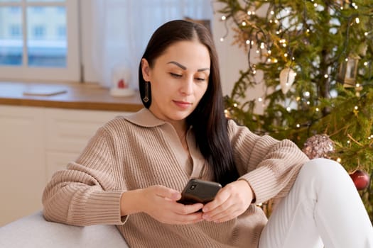 Woman with phone in hands sitting at home in kitchen on sofa during christmas holidays