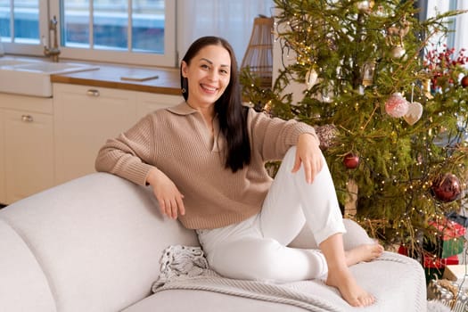 Happy woman sits on couch in sweater next to natural Christmas tree, holiday mood