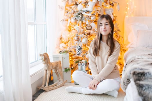 Cute girl sits on floor by window with Christmas tree in background in her bedroom