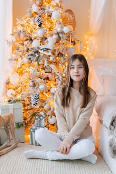Cute girl sits on floor by window with Christmas tree in background in her bedroom