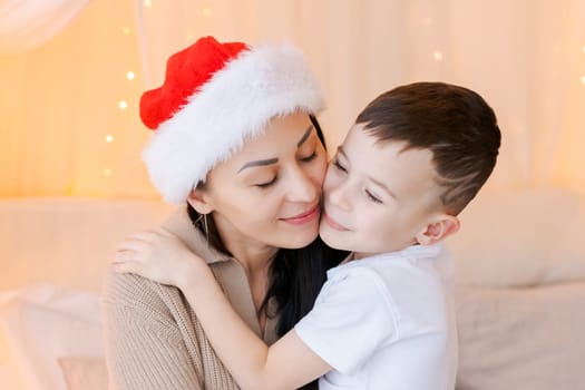 Happy mom and son wearing santa claus hat cuddle on bed in decorated bedroom on Christmas Eve in anticipation of the party