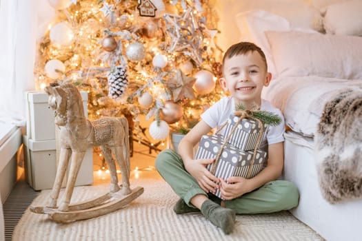 Happy boy sits on floor with gift in his hand by window in front Christmas tree in his bedroom