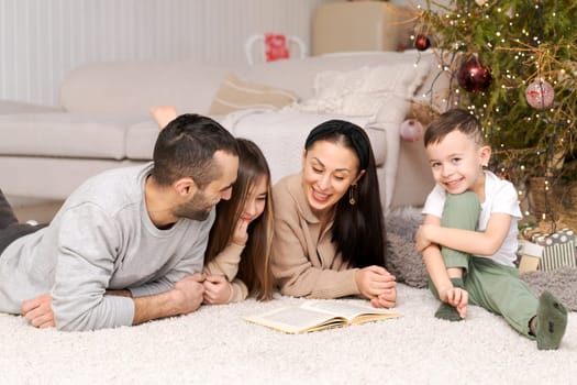 Happy family in decorated house at christmas lying on floor reading book against background of couch and Christmas tree. Selective focus