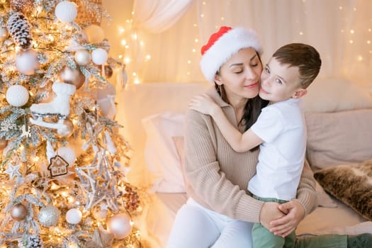 Happy mom and son wearing santa claus hat cuddle on bed in decorated bedroom on Christmas Eve in anticipation of the party