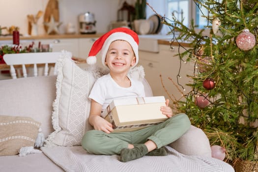 Cute little boy in Santa hat sits on couch with Christmas present from his family