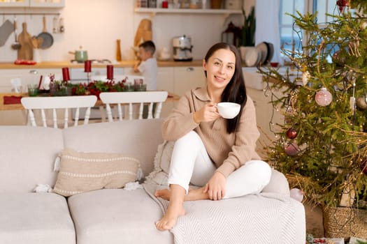 Concept festive Christmas atmosphere, lovely woman drinking tea or coffee sitting on a couch in a decorated kitchen wearing a sweater