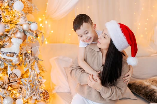 Happy mom and son wearing santa claus hat cuddle on bed in decorated bedroom on Christmas Eve in anticipation of the party