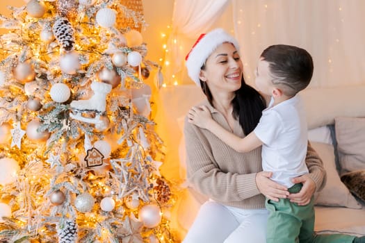 Happy mom and son wearing santa claus hat cuddle on bed in decorated bedroom on Christmas Eve in anticipation of the party