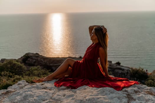 Woman sunset sea red dress, side view a happy beautiful sensual woman in a red long dress posing on a rock high above the sea on sunset