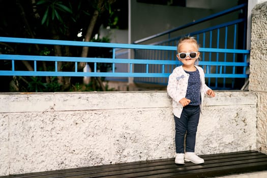 Little smiling girl in sunglasses stands on a bench near the garden fence. High quality photo