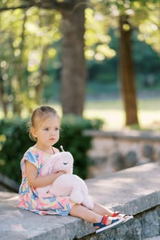 Little girl with a plush hare in her arms sits on a stone fence in the park. High quality photo