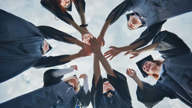 Team of college or university students celebrating graduation. Group of happy successful graduates in academic hats and robes standing in circle and putting their hands together.