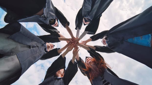 Team of college or university students celebrating graduation. Group of happy successful graduates in academic hats and robes standing in circle and putting their hands together.