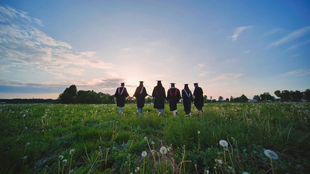 Six graduates in robes walk against the backdrop of the sunset