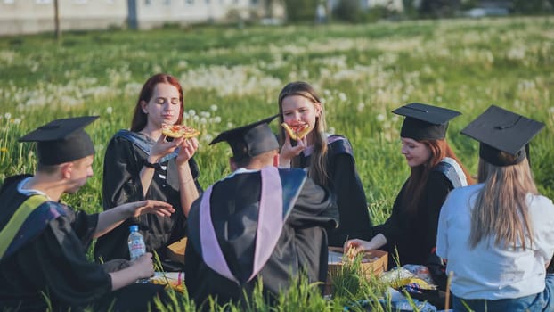 Graduates in black suits eating pizza in a city meadow