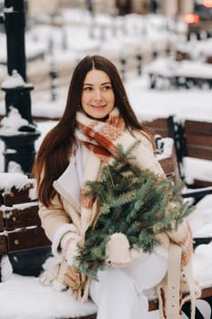 A girl with long hair in winter sits on a bench outside with a bouquet of fresh fir branches.