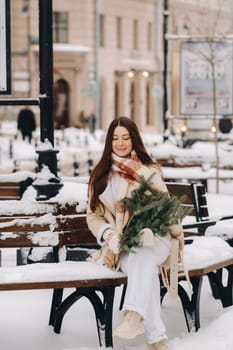 A girl with long hair in winter sits on a bench outside with a bouquet of fresh fir branches.