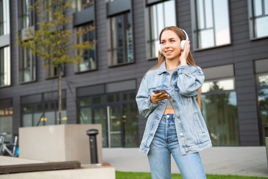 A cheerful young lady, sporting headphones and clutching a mobile device, beams with delight in the street