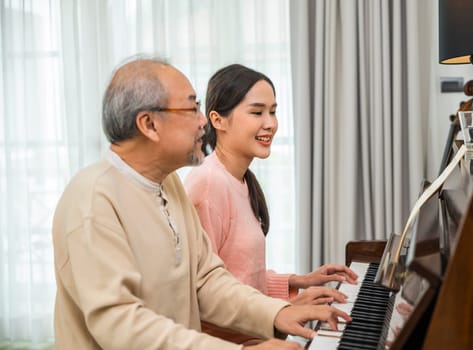 Family care. Asian young woman teaching piano for senior man teaching, happy daughter and elderly father playing the piano together in living room at home, lifestyle life after retirement