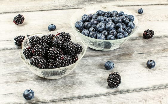 Blueberries and blackberries in glass bowls, some of them spilled on gray wood desk.