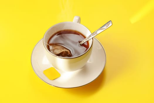 Morning sun shines on yellow table with white porcelain cup full of hot tea, bag still in