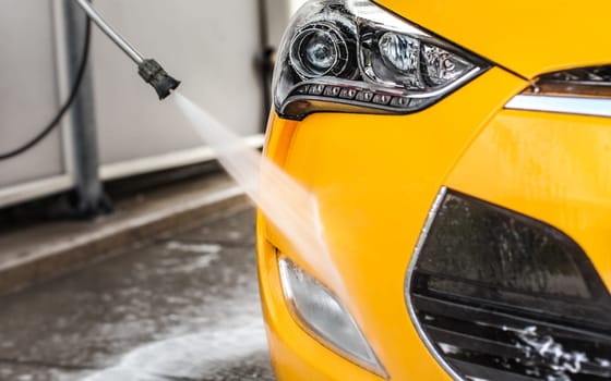 Front bumper and fog light of yellow car washed in self serve carwash; detail on jet water from nozzle spraying on glossy surface.