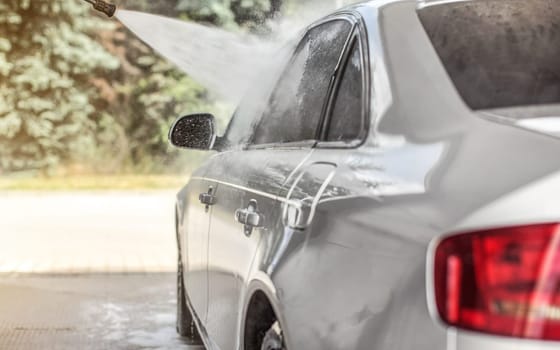 Silver car washed in self service carwash, jet water spraying from nozzle to side window, sun lit trees in background.