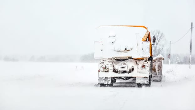 Orange plough truck on snow covered road, gray sky and trees in background, view from car driving behind