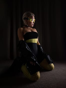 sexy girl in futuristic neon glasses and tight clothes and bright fishnet tights on a dark background in the room