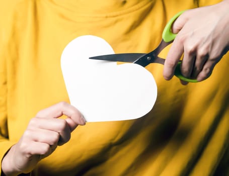 A girl holds scissors in her hands and cuts a heart, a young woman in a yellow sweater expresses her feelings, ends an abusive relationship with toxic love, dissolves a marriage, hands closeup.