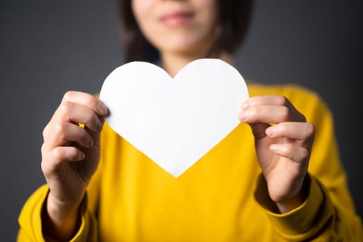 A young girl holds a white paper heart - a postcard or shows feelings of love. A woman with a joyful surprised face before the holiday and in search of love, relationships or shares her impressions.