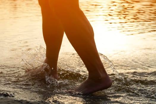A person runs along the beach on the water, the sea or ocean in the rays of the sun, an athlete man spends time happily, is training, jogging outdoors, close-up view of the legs and splashes.