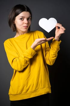 A young girl holds a white paper heart - a postcard or shows feelings of love. A woman with a joyful surprised face before the holiday and in search of love, relationships or shares her impressions.