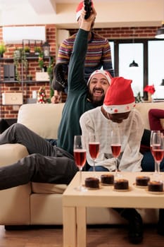 Happy man employee with joystick winning video game while celebrating christmas with colleagues in office. Excited company worker holding gamepad while enjoying new year corporate party